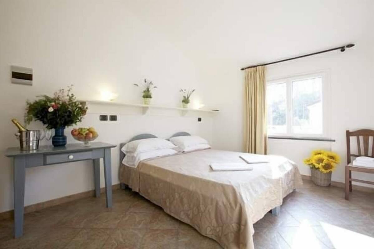 Ein Schlafzimmer mit einem Bett, einem Schreibtisch mit Blumenvase und einem Fenster mit Vorhängen.