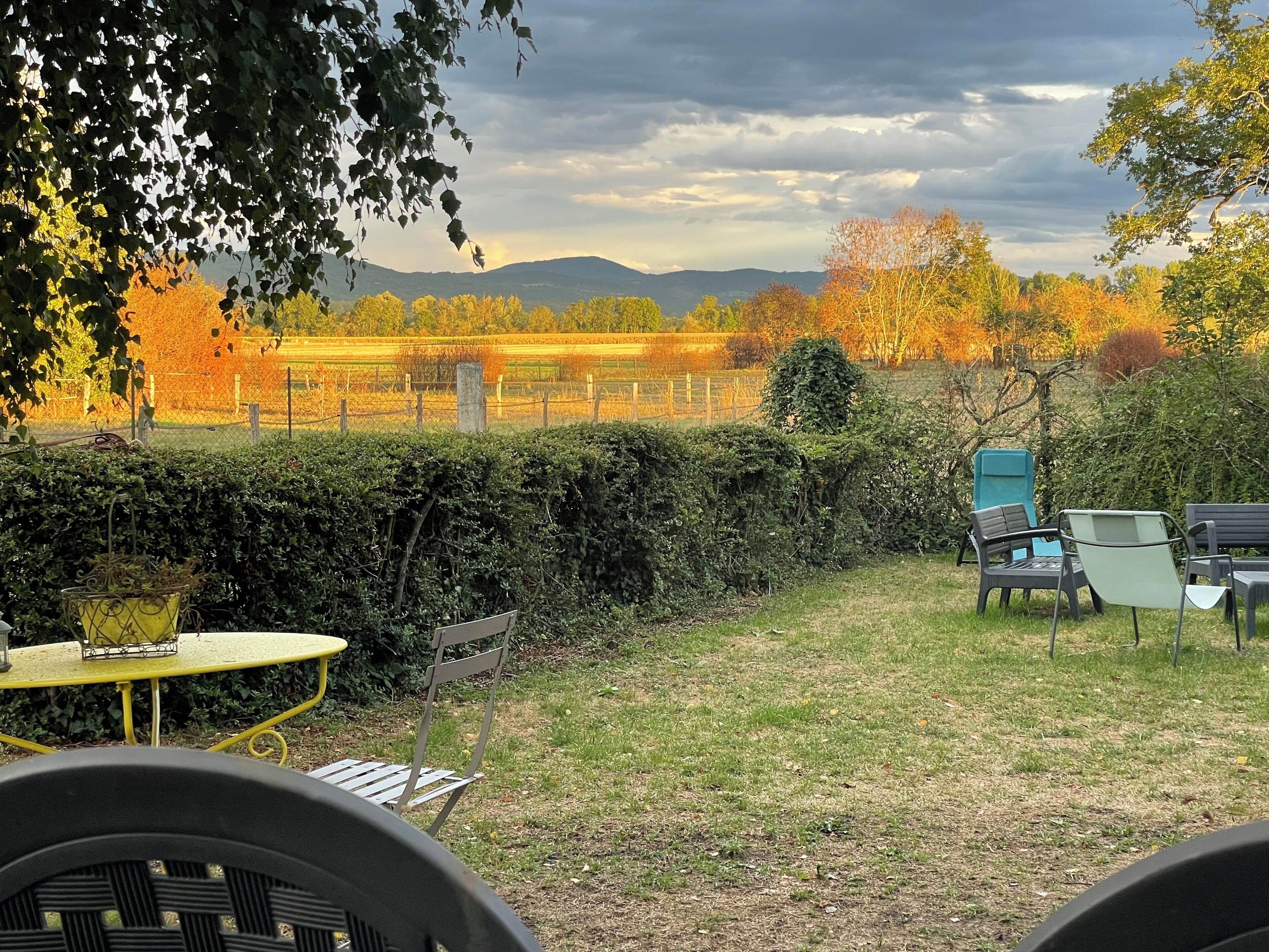 Un jardin avec une table et des chaises, une haie, et une vue sur des champs et des collines.