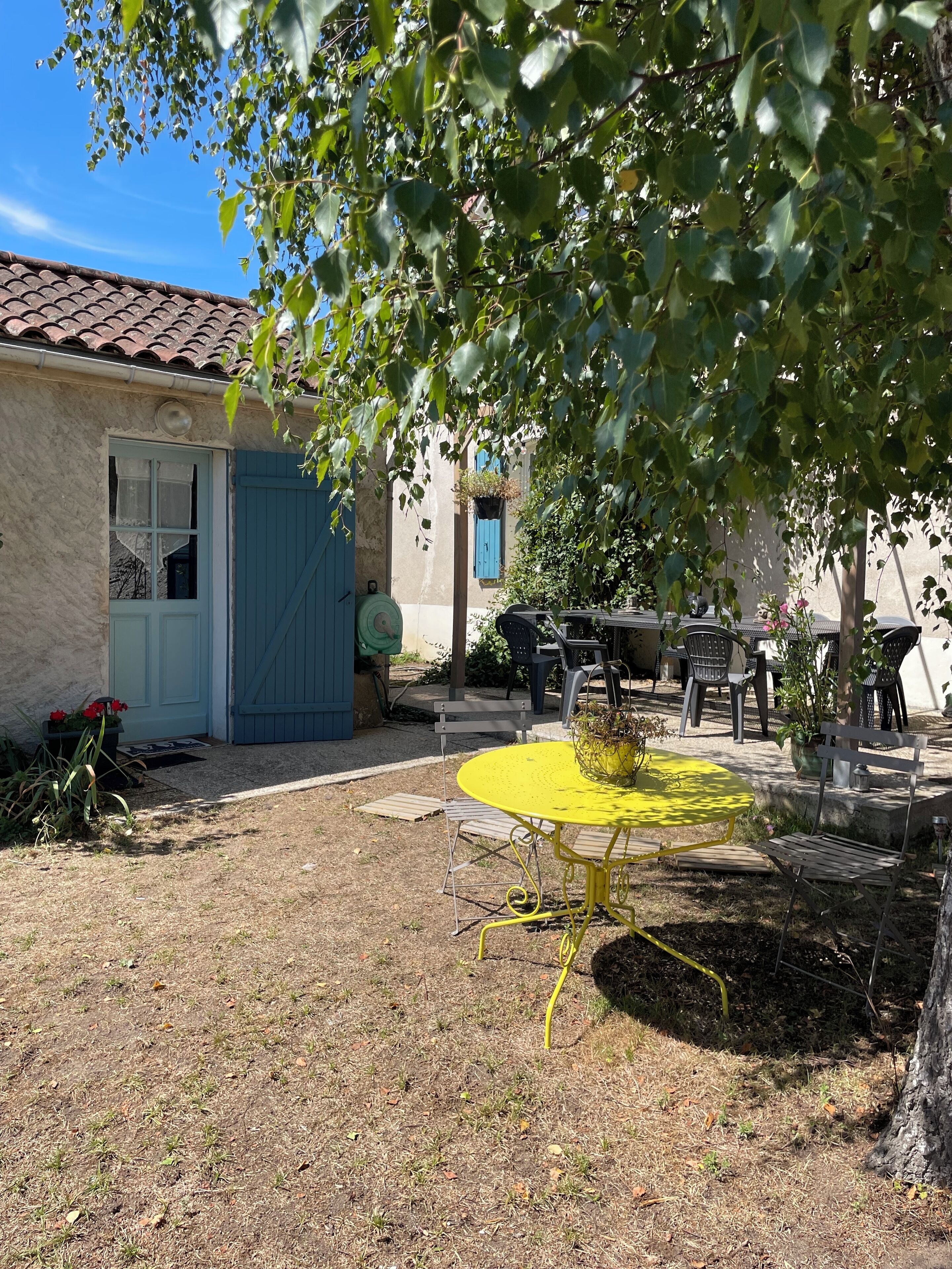 A rustic outdoor seating area with a yellow metal table and chairs, surrounded by greenery and a building with a blue door and windows.