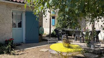 Un espace extérieur aménagé dans un style rustique, avec une table et des chaises en métal jaune, entouré de verdure et bordé d’un bâtiment doté d’une porte et de fenêtres bleues.