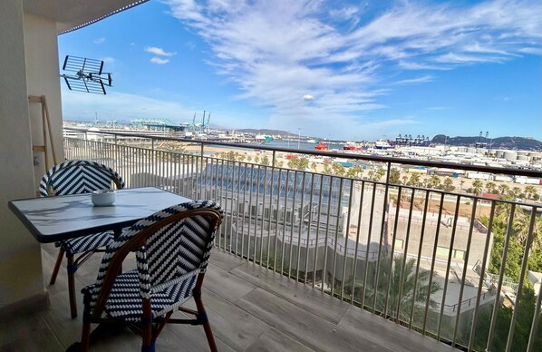 A balcony with a table and chairs, overlooking a marina with boats and industrial structures. - Modern, spacious apartment in Algeciras | Bay view | Near the port (Algeciras)