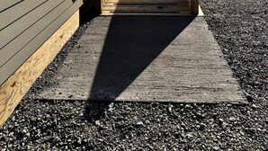 Una escalera de madera que conduce a la entrada de un edificio, rodeada de grava y árboles.