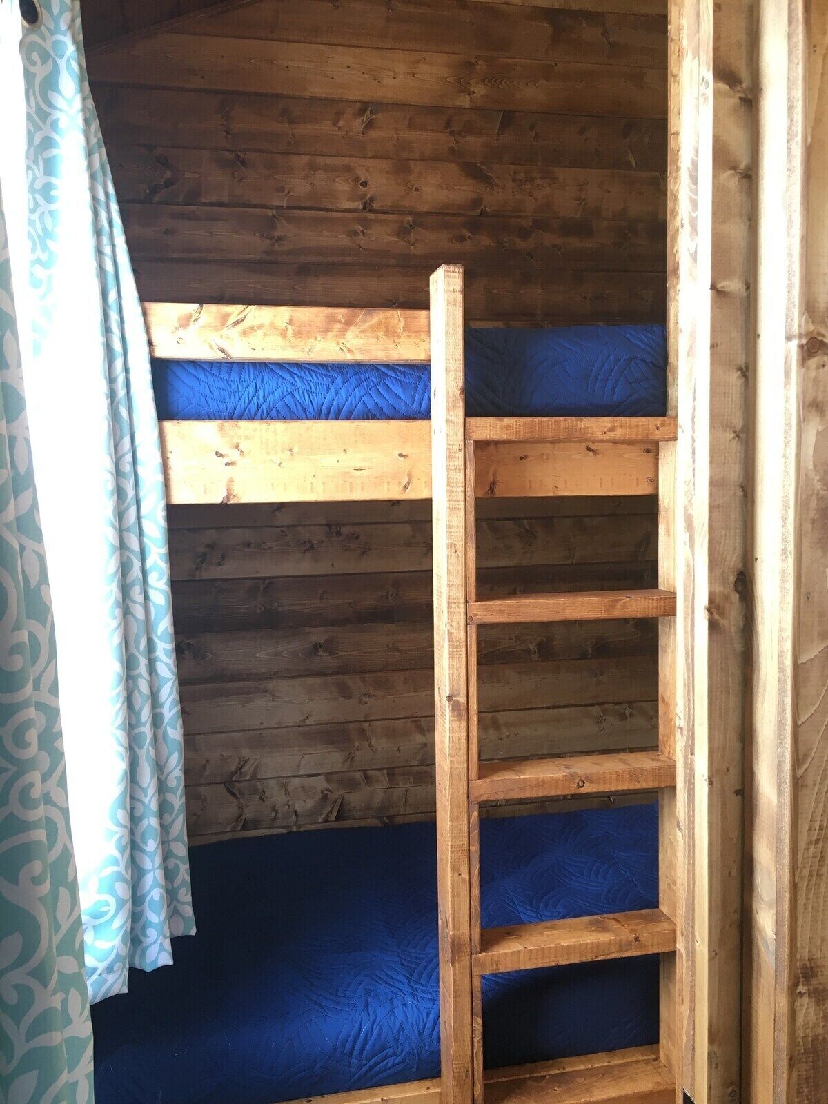Una cama litera con escalera de madera, ropa de cama azul y cortina estampada.