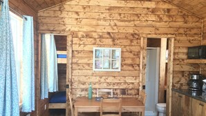 Interior de una cabaña de madera con una mesa de comedor, sillas, una pequeña cocina con microondas y una ventana con cortinas.