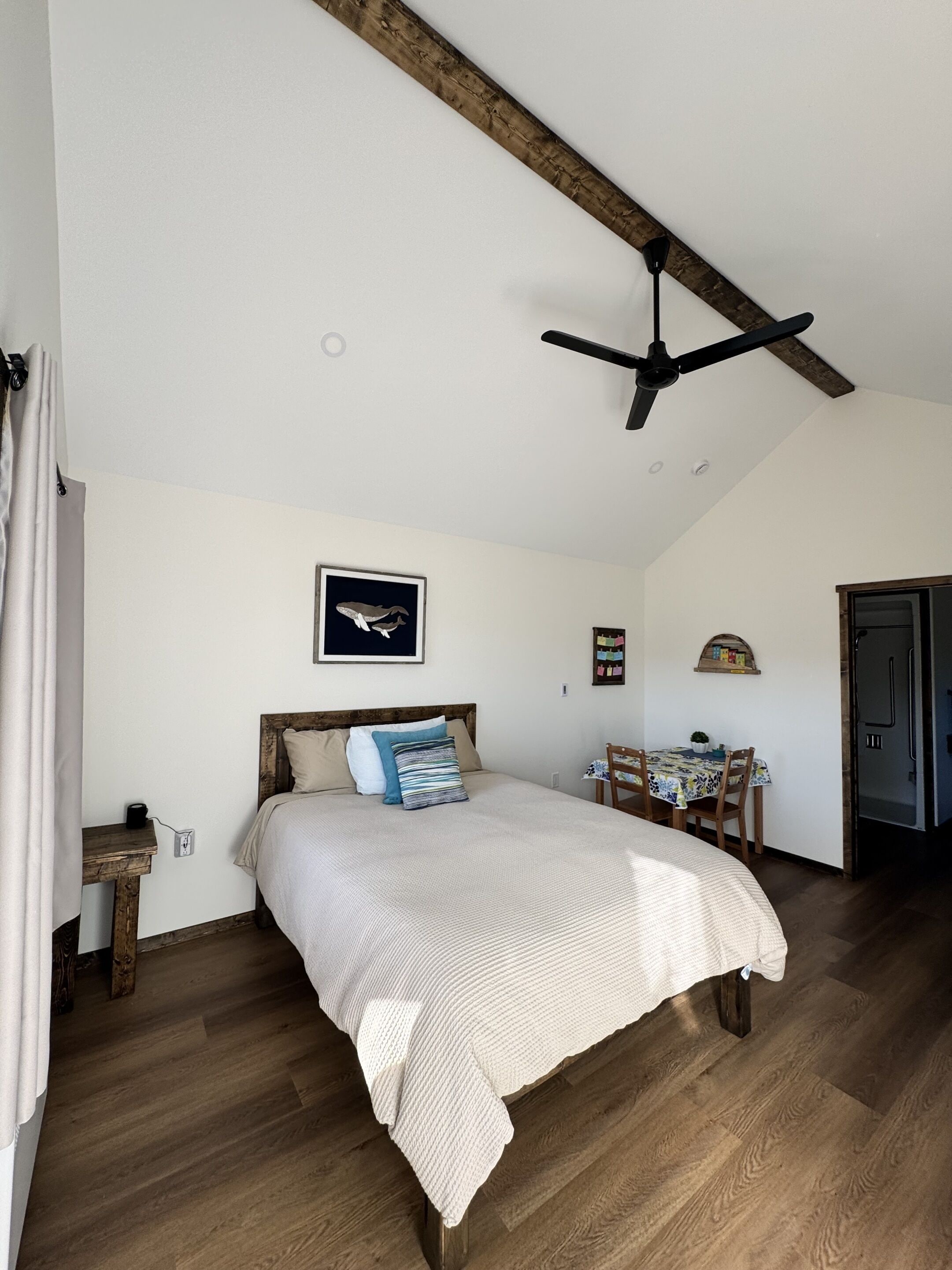 A bedroom with a bed, wooden floor, ceiling fan, and a dining area with a table and chairs.