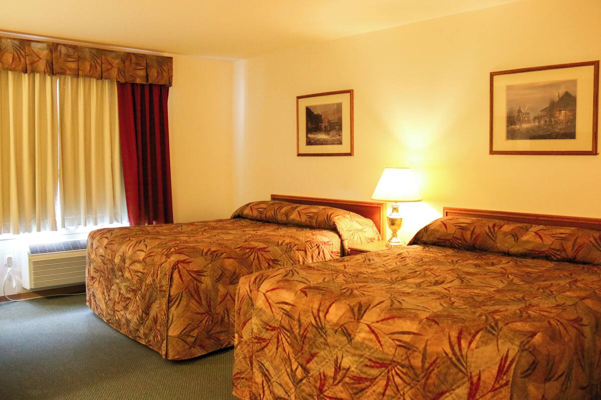 A hotel room with two beds, a window with curtains, a wall-mounted lamp, and two framed pictures.