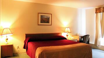 A hotel room with a bed, two bedside lamps, a desk with a chair, and a window with curtains.
