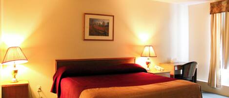 Habitación de hotel con cama, dos lámparas de noche, un escritorio con silla y una ventana con cortinas.