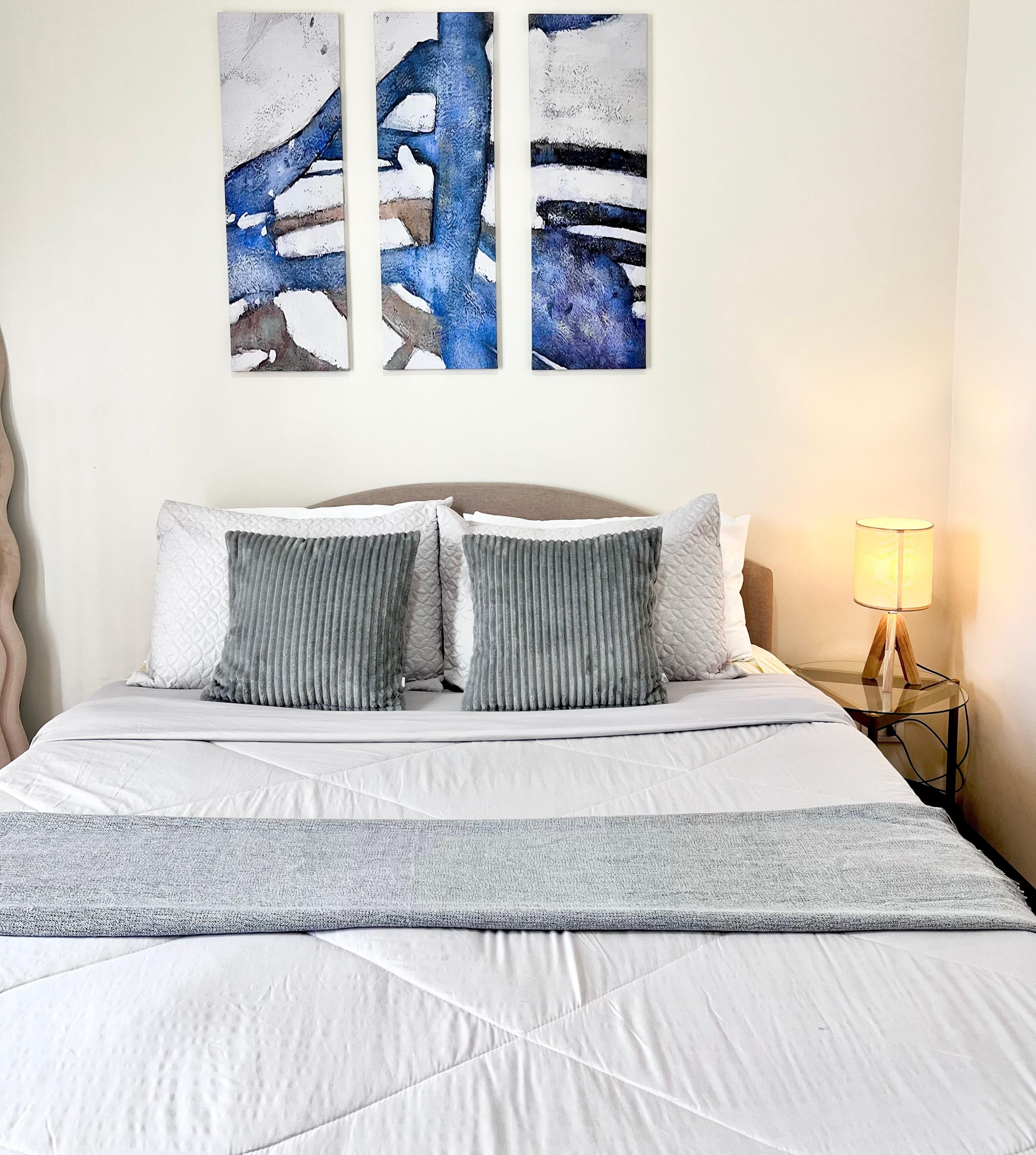 A bed with a white quilt, grey pillows, and a grey throw blanket. There are three abstract paintings on the wall above the bed.