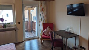 A room with a wooden ceiling, a TV mounted on the wall, a red chair, a table, and a view through a glass door to an outdoor balcony. - Gasthaus zum Hirschen (Muhr am See)