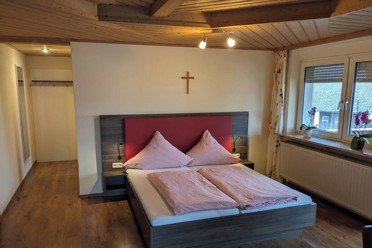 Une chambre avec un plancher en bois, un lit doté d’une tête de lit rouge, une croix accrochée au mur et une fenêtre avec des rideaux.