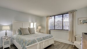 A bedroom with a bed, bedside tables, a chair, a window with curtains, and a painting on the wall. - 4205 Island Club - Folly Field - 2 Bed 2 Bath Sleeps 5 - Hilton Head Rentals (Hilton Head Island)