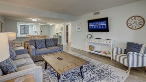 A modern living room with a sofa, a coffee table, a TV mounted on the wall, and a clock. - 4205 Island Club - Folly Field - 2 Bed 2 Bath Sleeps 5 - Hilton Head Rentals (Hilton Head Island)