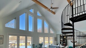 A spacious living room with a high ceiling, large windows, a ceiling fan, a spiral staircase, and a cozy seating area.