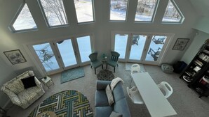 A spacious living room with a high ceiling, large windows, a grey sofa, a white table, and a bookshelf.