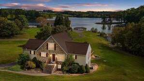 A stone house with a wrap-around porch, a well-maintained lawn, and a view of a lake and distant buildings.