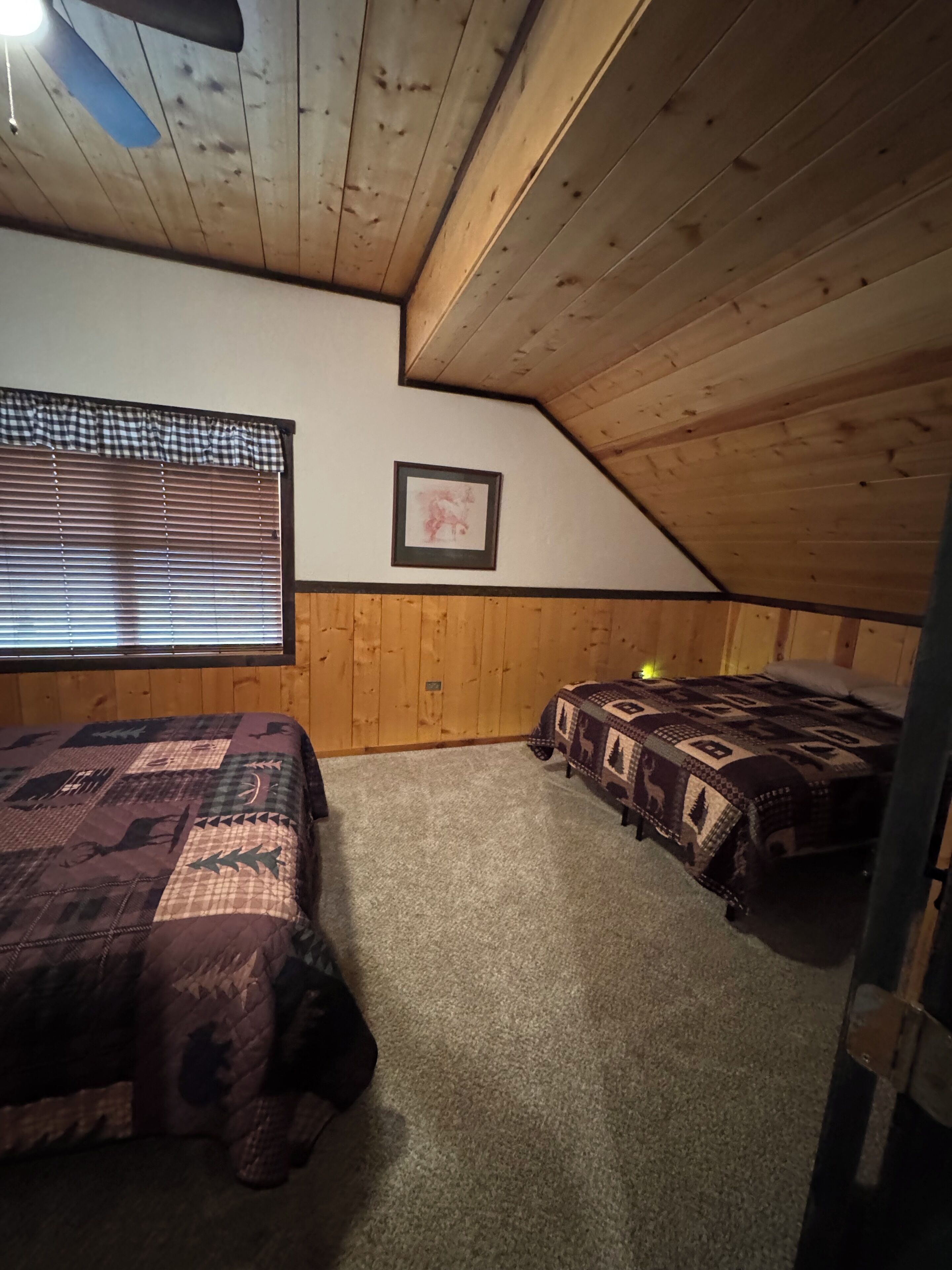 Une chambre confortable située sous les toits, avec deux lits, des murs en bois, un tableau encadré et une fenêtre avec des stores.