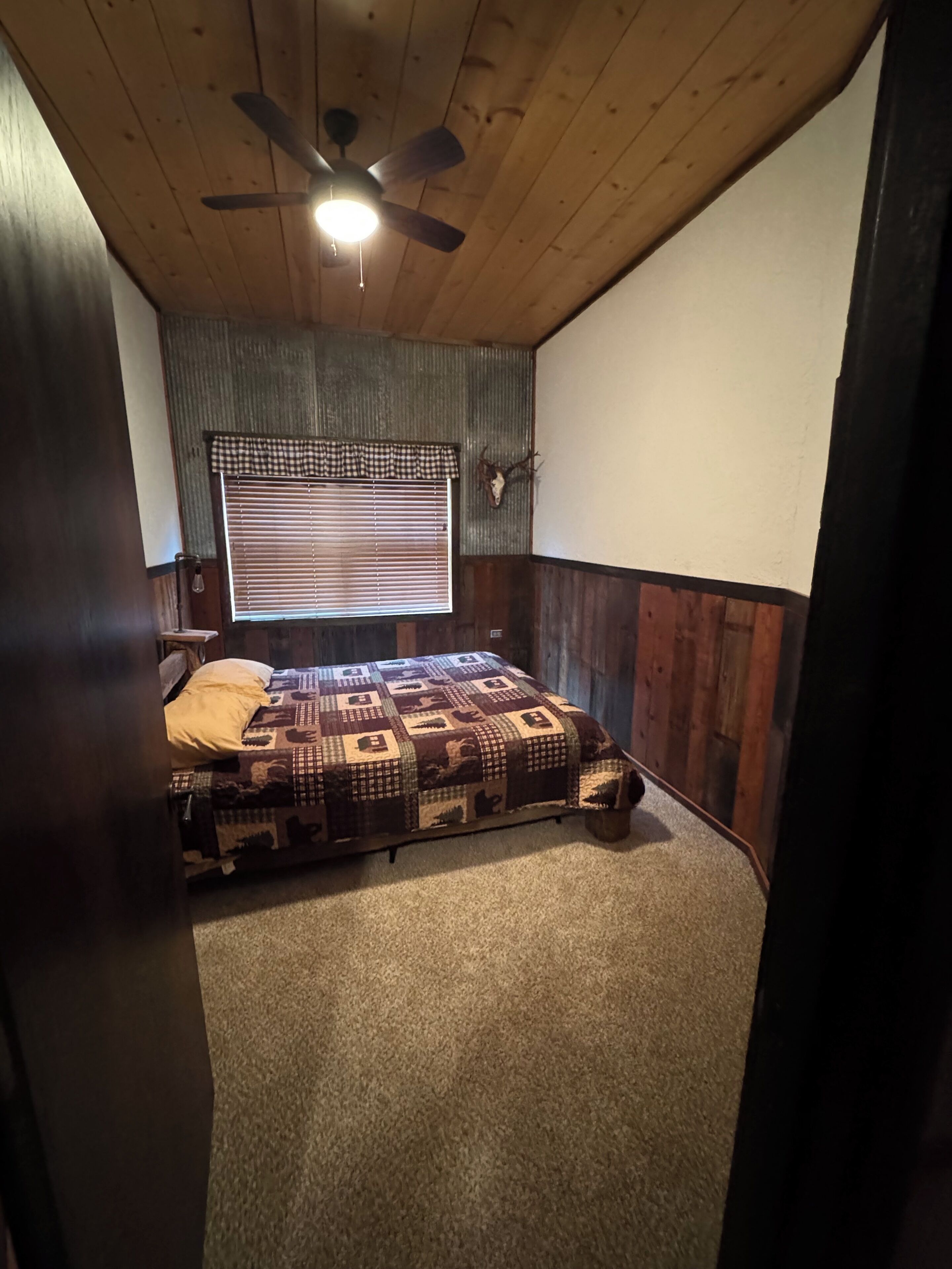 A bedroom with a bed, a ceiling fan, a window with blinds, and a wall-mounted deer skull.
