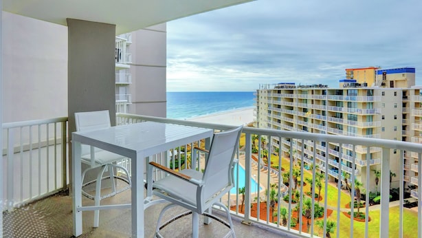 Un balcón con una mesa y una silla blancos, con vistas a una piscina y una playa.