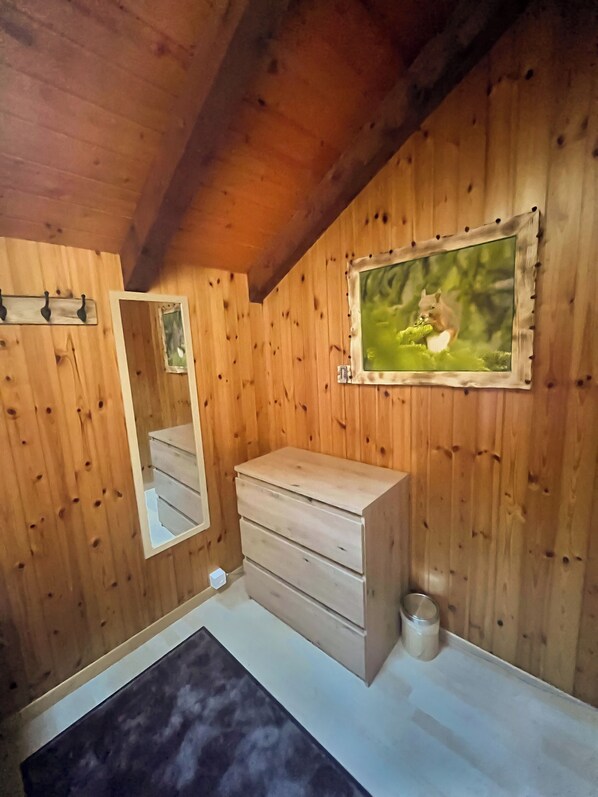 Een kamer met houten lambrisering, een houten dressoir, een spiegel, een lijst met een foto en een kleine prullenbak.