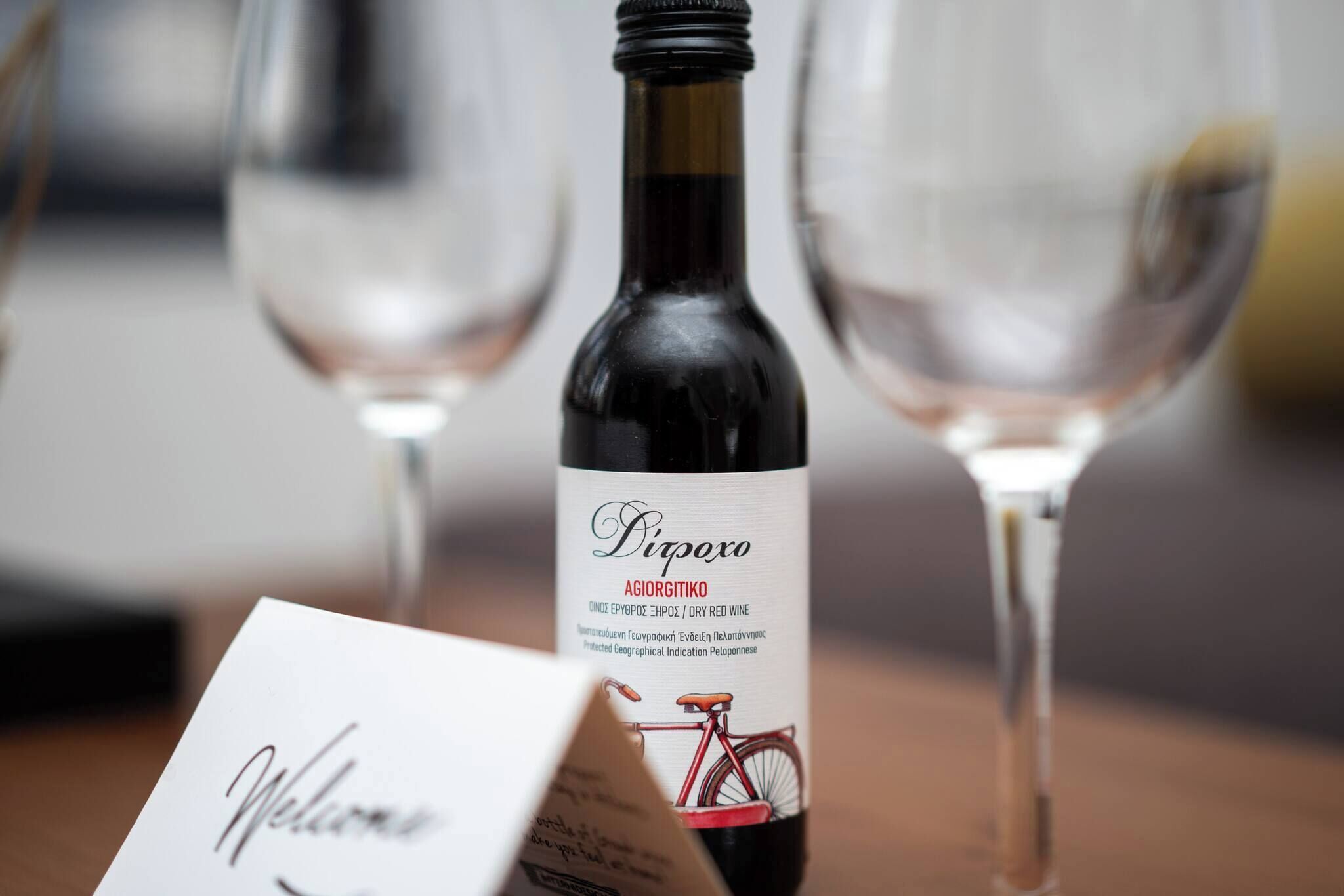 Uma garrafa de vinho Agiorgitiko com rótulo que apresenta uma ilustração de bicicleta, colocada sobre uma mesa com duas taças de vinho.