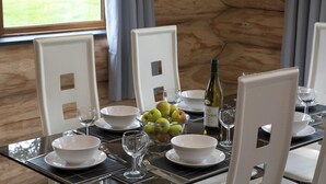 A dining table set with white plates, bowls, and a bowl of green apples. There are wine glasses and a bottle of wine on the table. The room has wooden walls and a window with curtains. - Traditional Scandinavian/north American Log Home (Ripon)