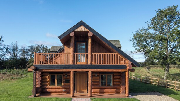 A log cabin with a balcony, a front door, and a gravel driveway. - Traditional Scandinavian/north American Log Home (Ripon)