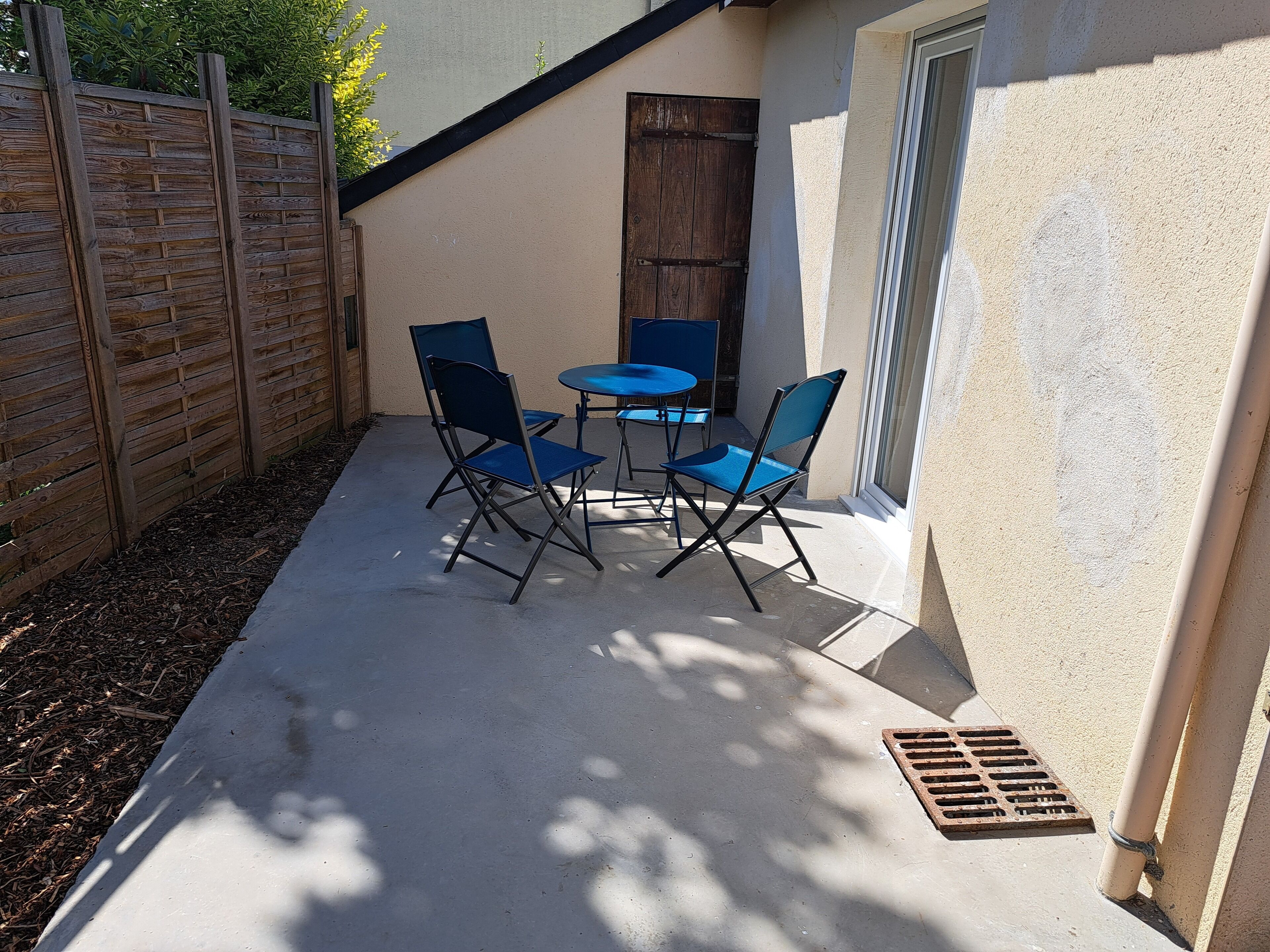Een terras buiten met een betonnen vloer, een houten omheining en een blauwe tafel met vier stoelen.
