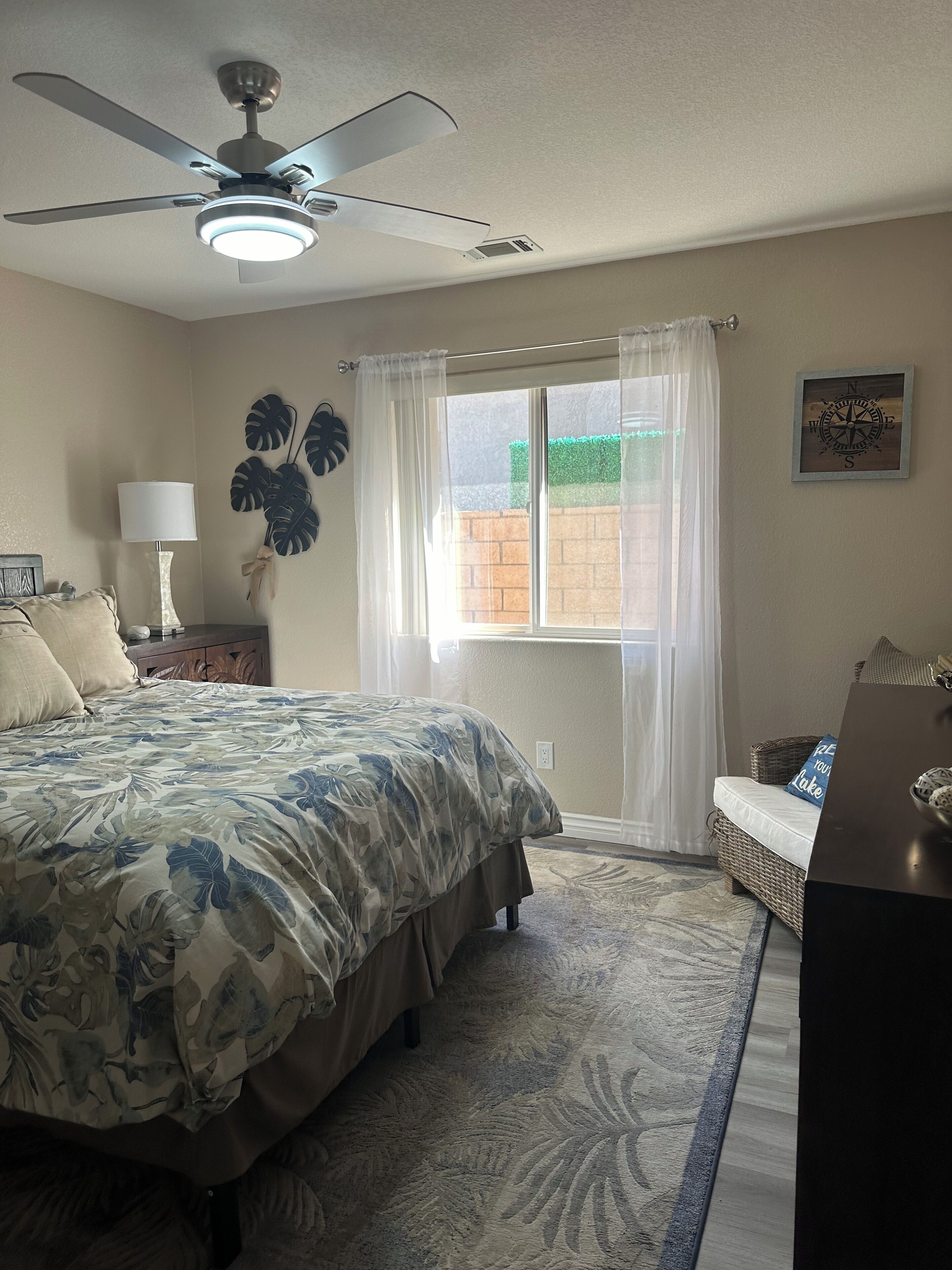 A bedroom with a bed, a ceiling fan, a window with curtains, a bedside table with a lamp, and a wall clock.