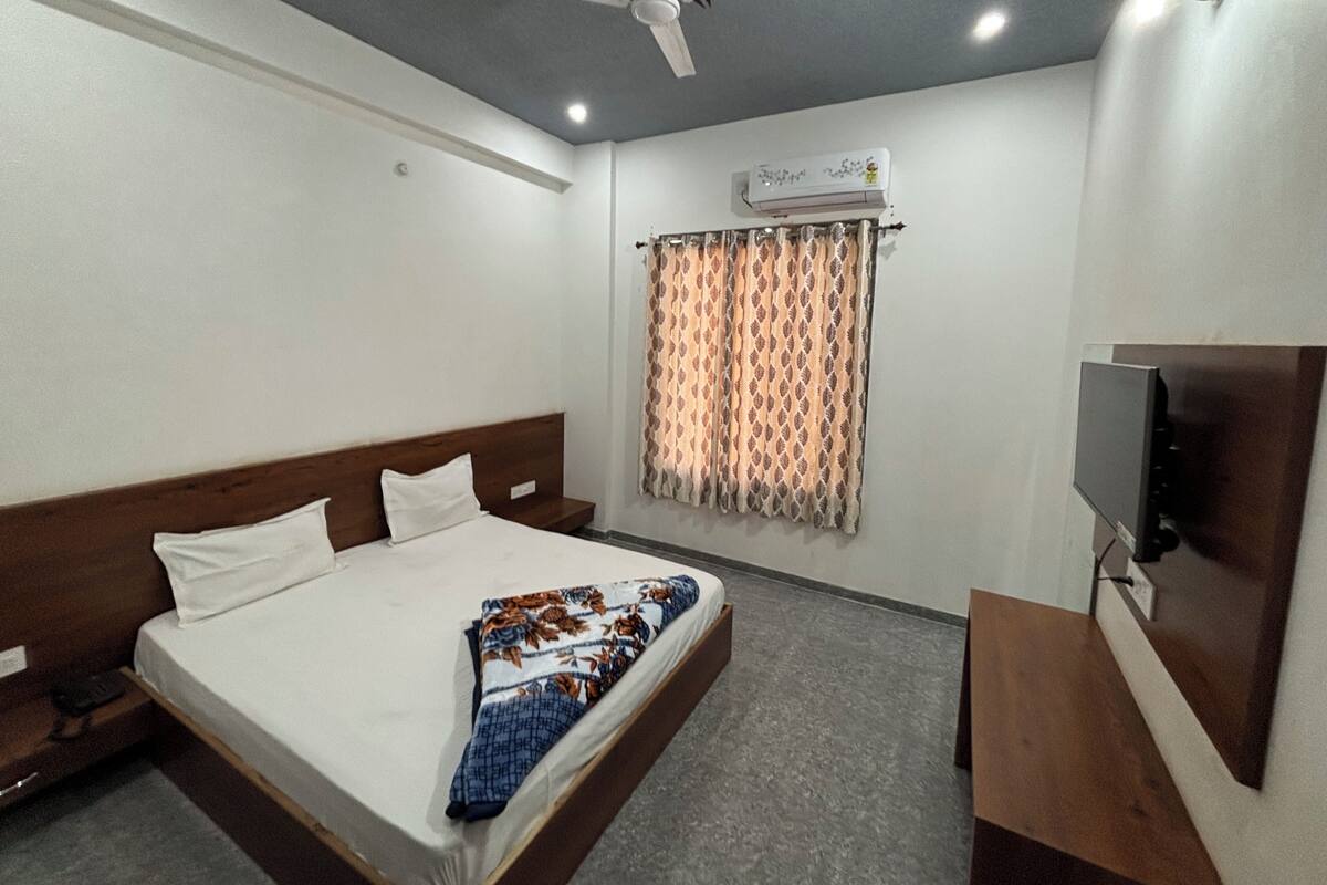 Ein Hotelzimmer mit einem Einzelbett, einem Holz-Kopfteil, einem Deckenventilator, einem Fenster mit gemusterten Vorhängen und einem an der Wand montierten Flachbildfernseher.