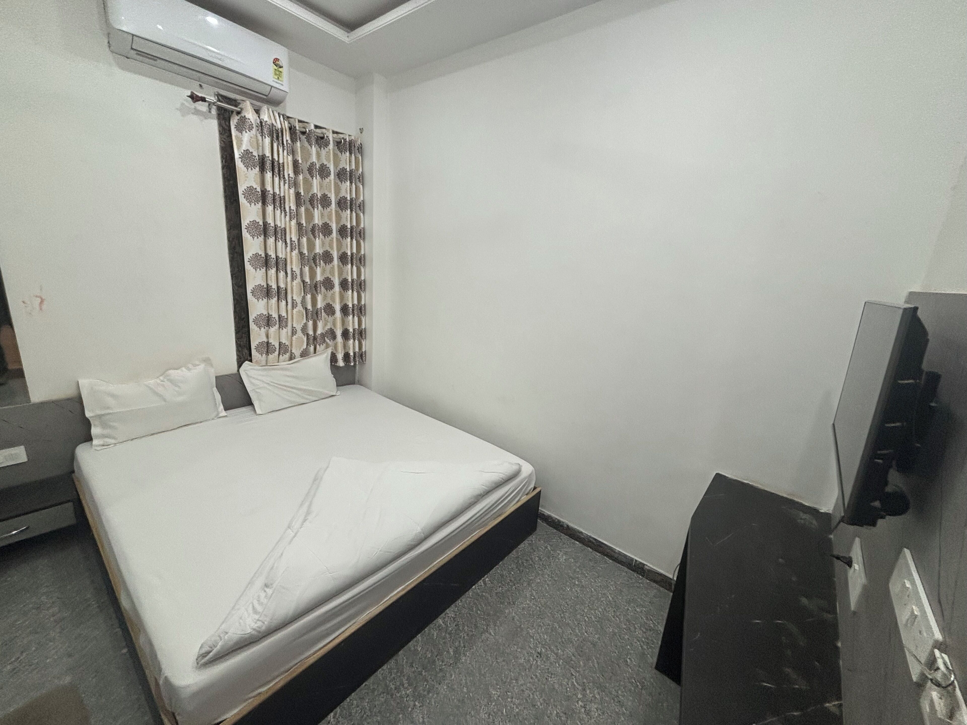 Hotellihuone, jossa on sänky, taulutelevisio, ilmastointilaite ja kukkakuviollinen verho.