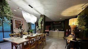 Ένας χώρος τραπεζαρίας με ένα μεγάλο ξύλινο τραπέζι, καρέκλες και ένα πολυέλαιο. Στο βάθος, υπάρχουν γλάστρες με φυτά και μια μικρή κουζίνα.