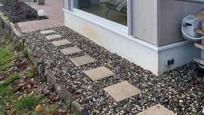 A modern building with a large window, a concrete exterior, and a gravel pathway leading to a door.