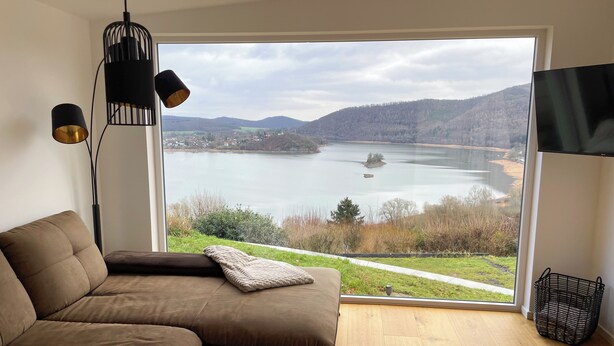 A modern living room with a large window offering a view of a lake and hills.
