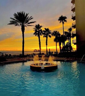 A swimming pool with a water fountain at sunset, surrounded by palm trees and a building. - Stunning Views! Shores of Panama #1216 (Panama City Beach)