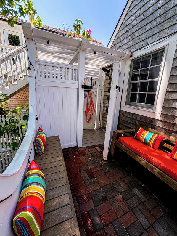A covered patio area with a wooden bench and colorful cushions, a white fence, and a window with blinds.