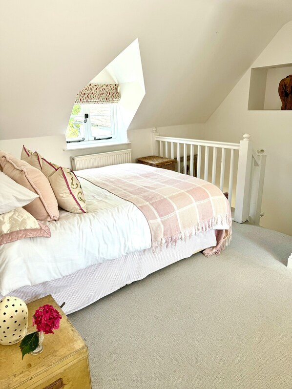 A bedroom with a bed, a window, a white railing, and a small wooden table with a vase of flowers. - Perfect Countryside Cottage (Fyfield)