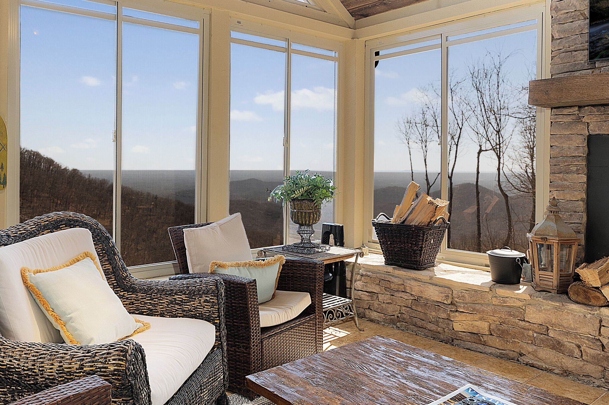 A sunlit room with wicker furniture, a stone fireplace, and large windows overlooking a mountainous landscape.