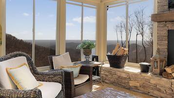 A sunlit room with wicker furniture, a stone fireplace, and large windows overlooking a mountainous landscape.