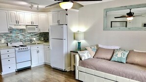 Une cuisine compacte avec des meubles blancs, un réfrigérateur, une cuisinière et un four à micro-ondes. On y trouve un lit avec une couverture matelassée et des oreillers, et un ventilateur de plafond au-dessus.