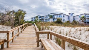 Une promenade en bois mène à un hôtel en bord de mer, doté de nombreux balcons et donnant sur une plage de sable.