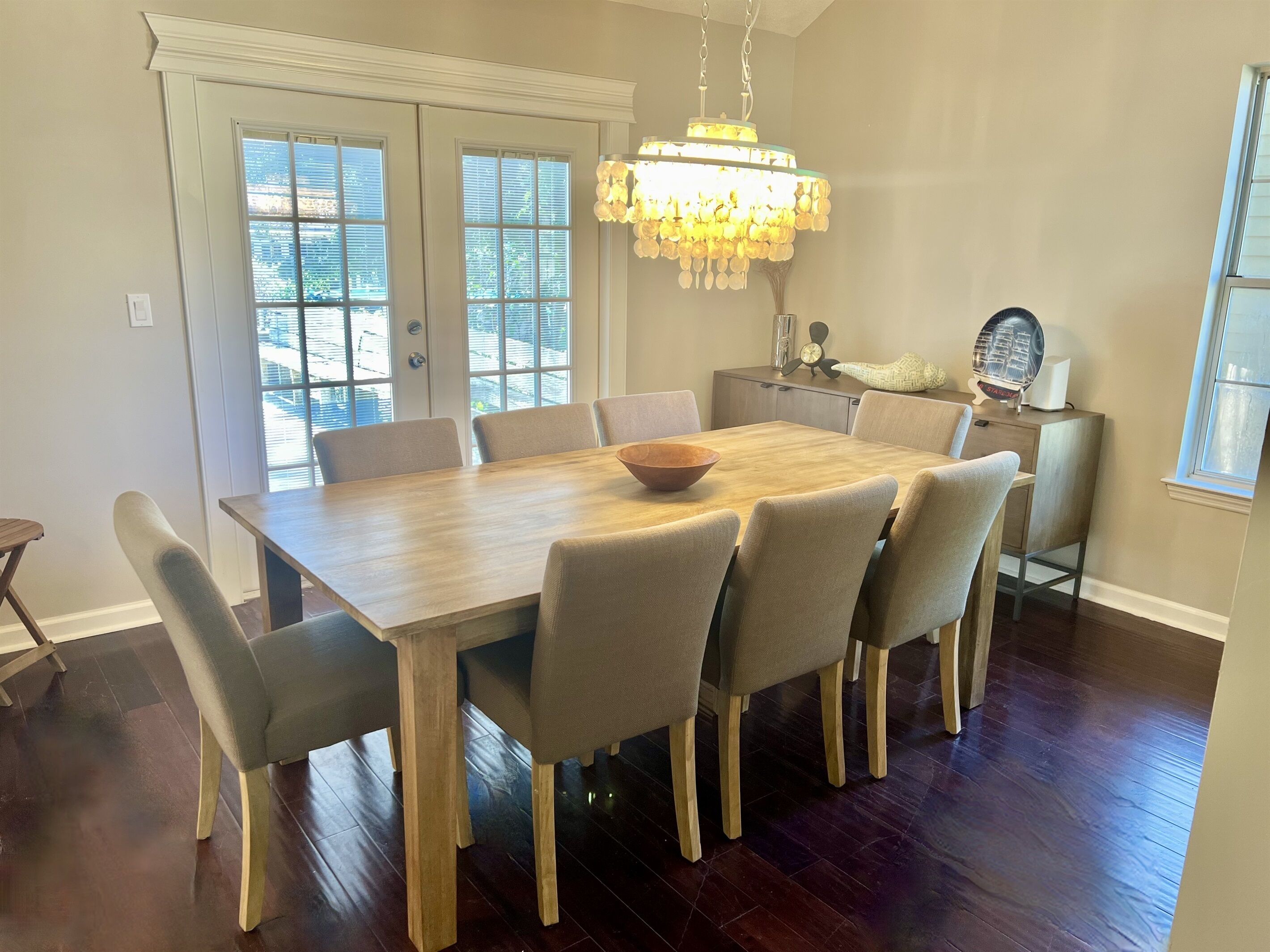 A dining area with a wooden table, six chairs, a chandelier, and a view of the outdoors through large windows.