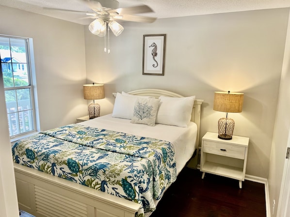 A bedroom with a bed, two bedside tables, a ceiling fan, a window, and a framed seahorse artwork.
