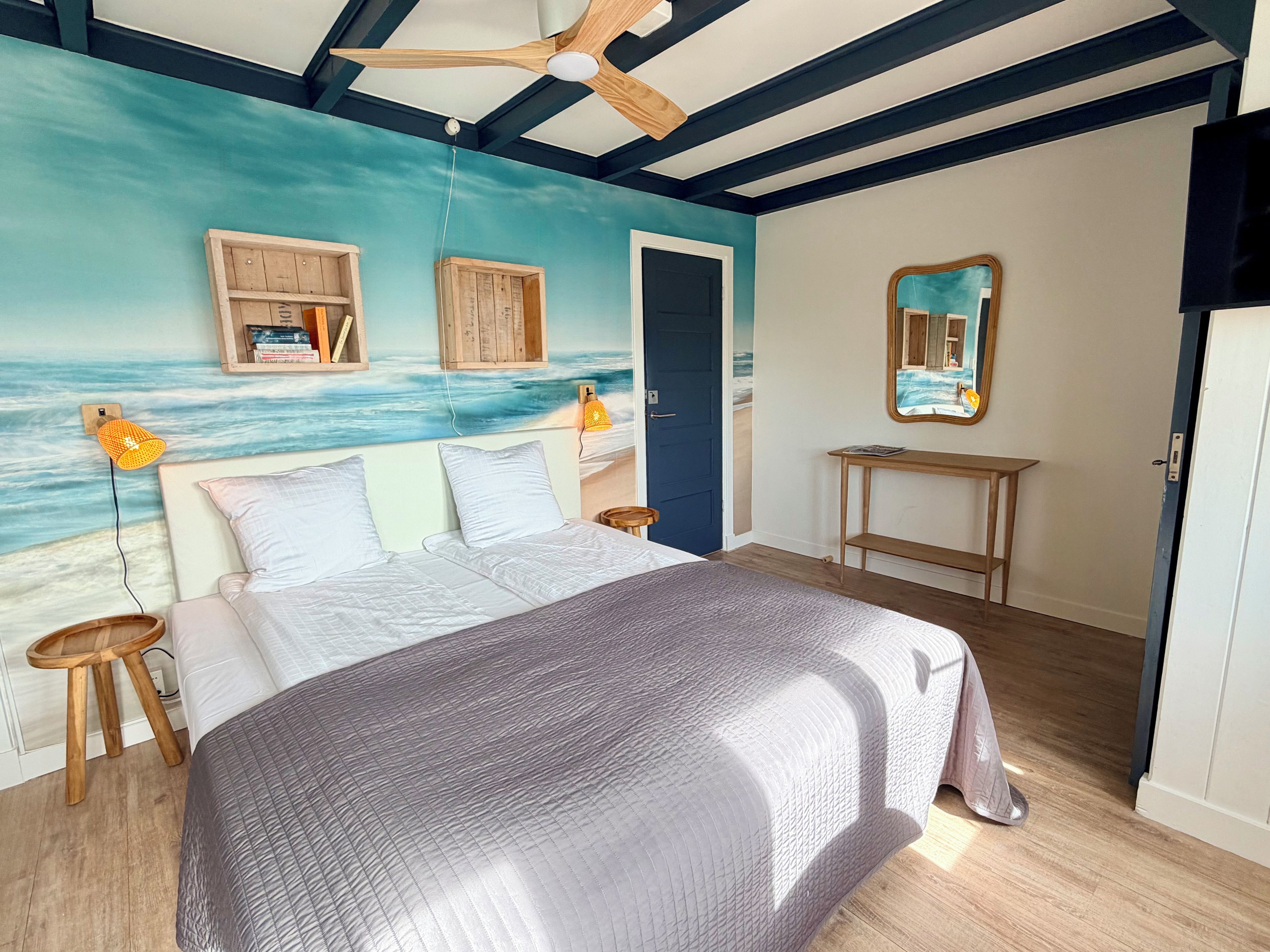 Un dormitorio con cama, cabecera de madera, un mural en la pared con playa, mesita de noche de madera, lámpara de noche y una puerta.