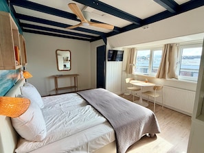 Une chambre à coucher comprenant un lit, un bureau, une chaise, une fenêtre avec des rideaux et un ventilateur de plafond.