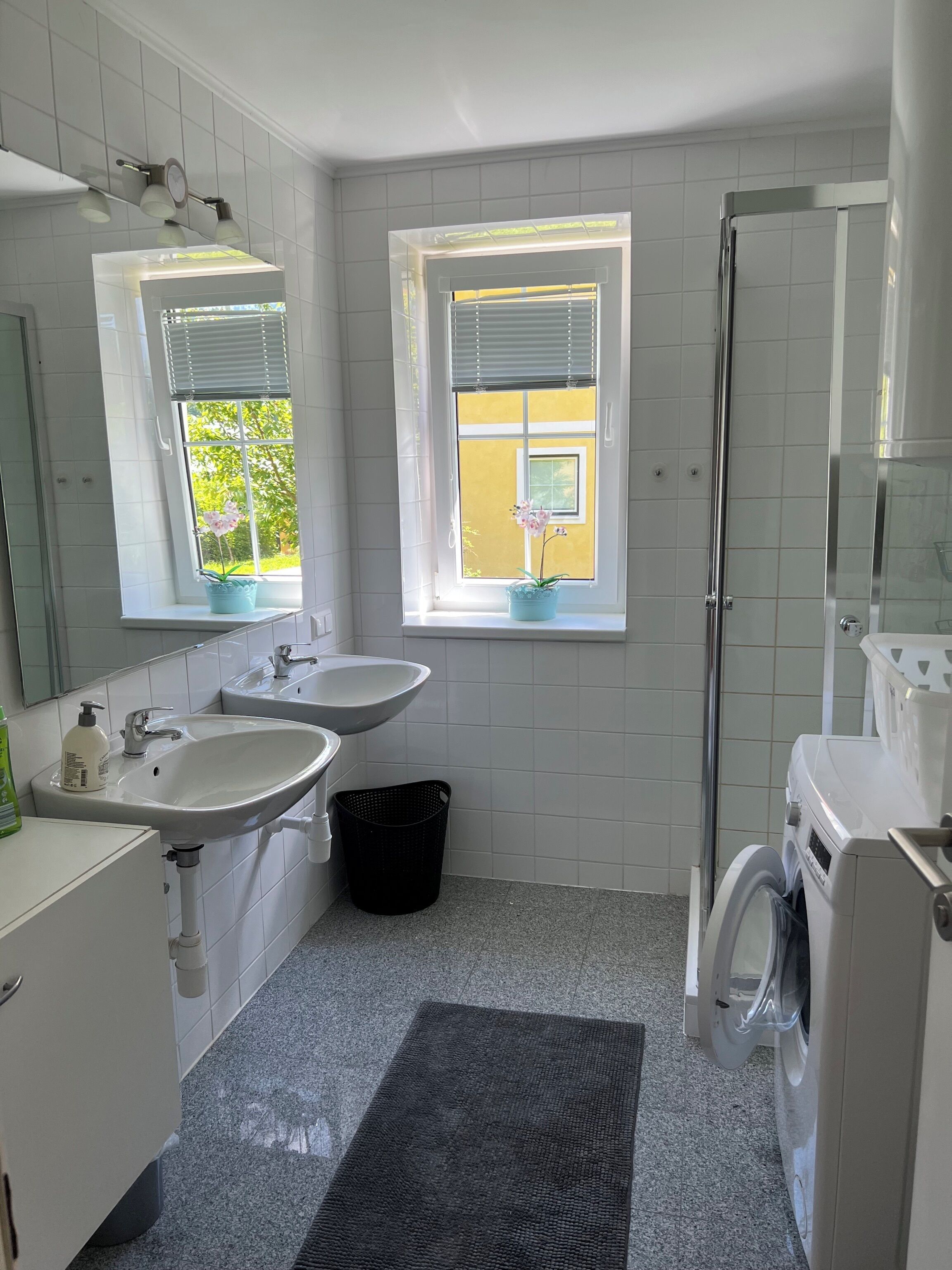 Un baño con dos lavamanos, un espejo, una ventana con persianas, una ducha y una lavadora.