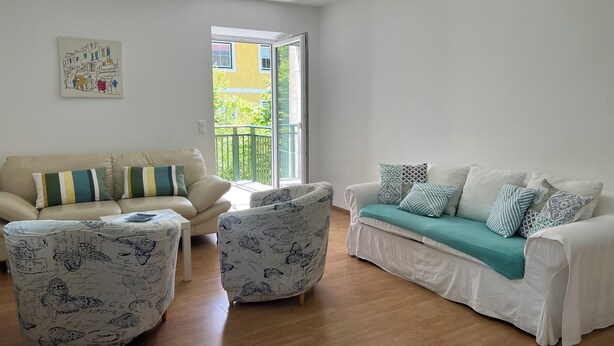 A living room with two sofas, a coffee table, and patterned armchairs. A large window provides a view of a balcony and greenery outside.