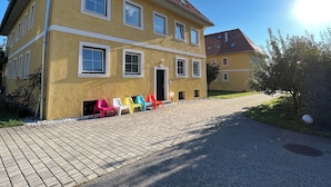 A yellow building with multiple windows and a red roof. There are colorful chairs lined up in front of the building.
