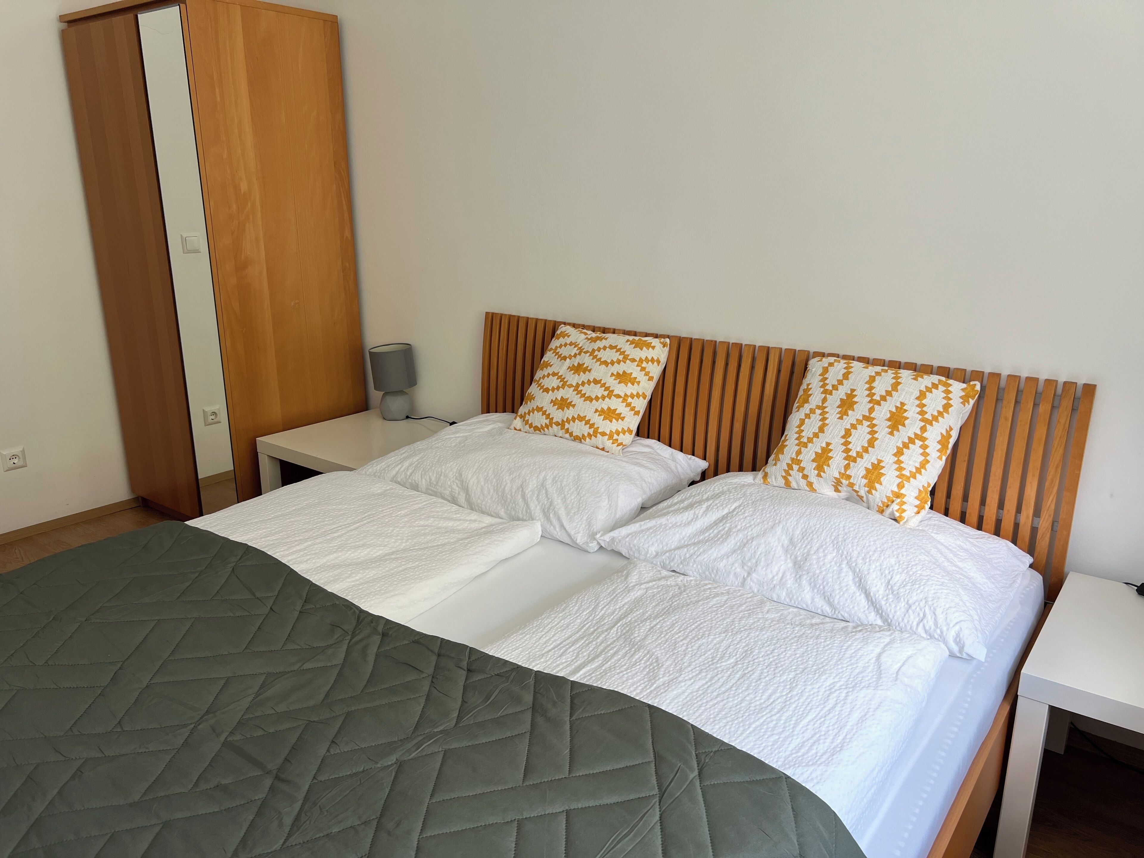 Ein Doppelbett mit einer grünen Tagesdecke, zwei Kissen mit geometrischen Mustern, ein Holz-Kopfbrett, ein Nachttisch mit Lampe und ein Kleiderschrank mit Spiegel.