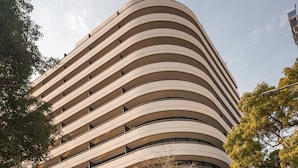 Um edifício com várias andares e fachada curva, área externa com mesas e plantas em vasos, e uma vitrine com sinalização.